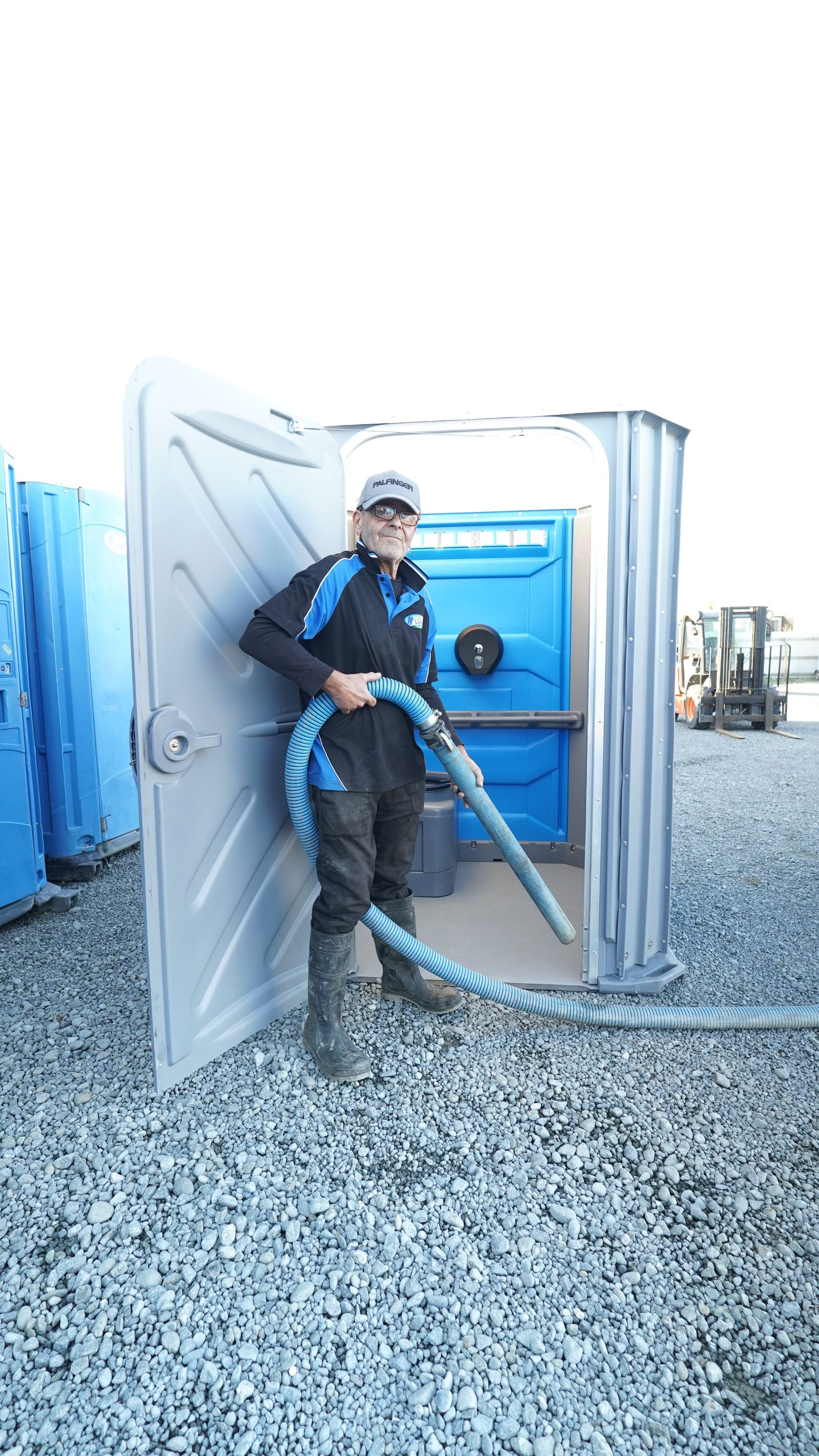 Man in uniform with hose cleaning a blue portable toilet. Gravel ground, other toilets visible.