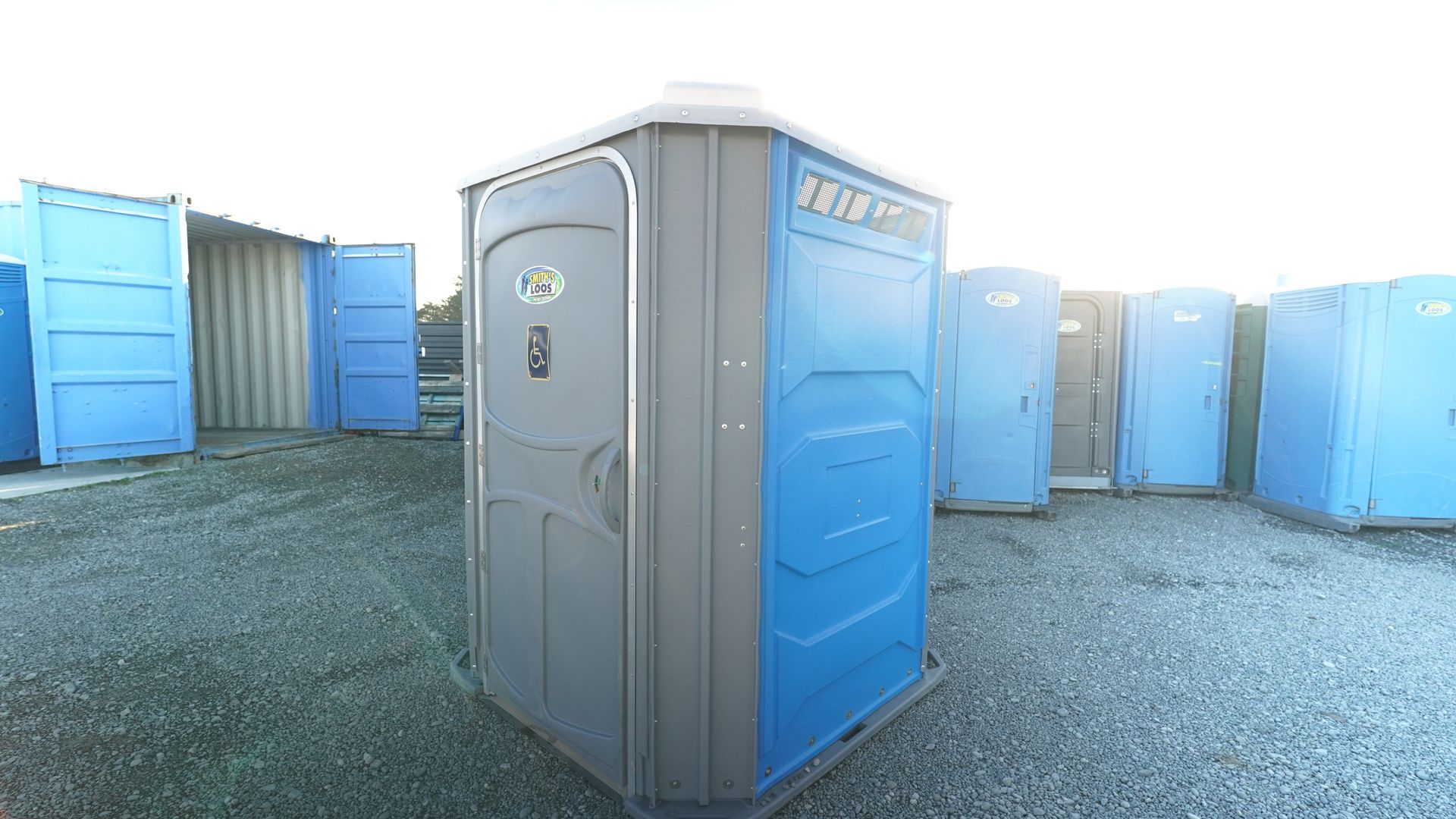 A gray and blue portable toilet stands among other blue portable toilets on gravel.