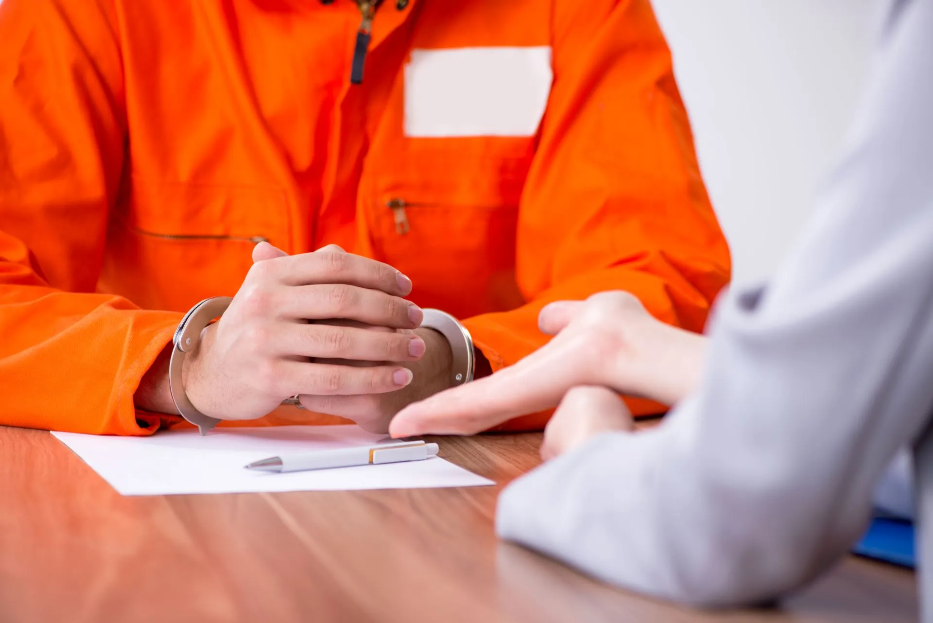 A person in orange prison jumpsuit with handcuffs sits at a table. Another person points at a paper.