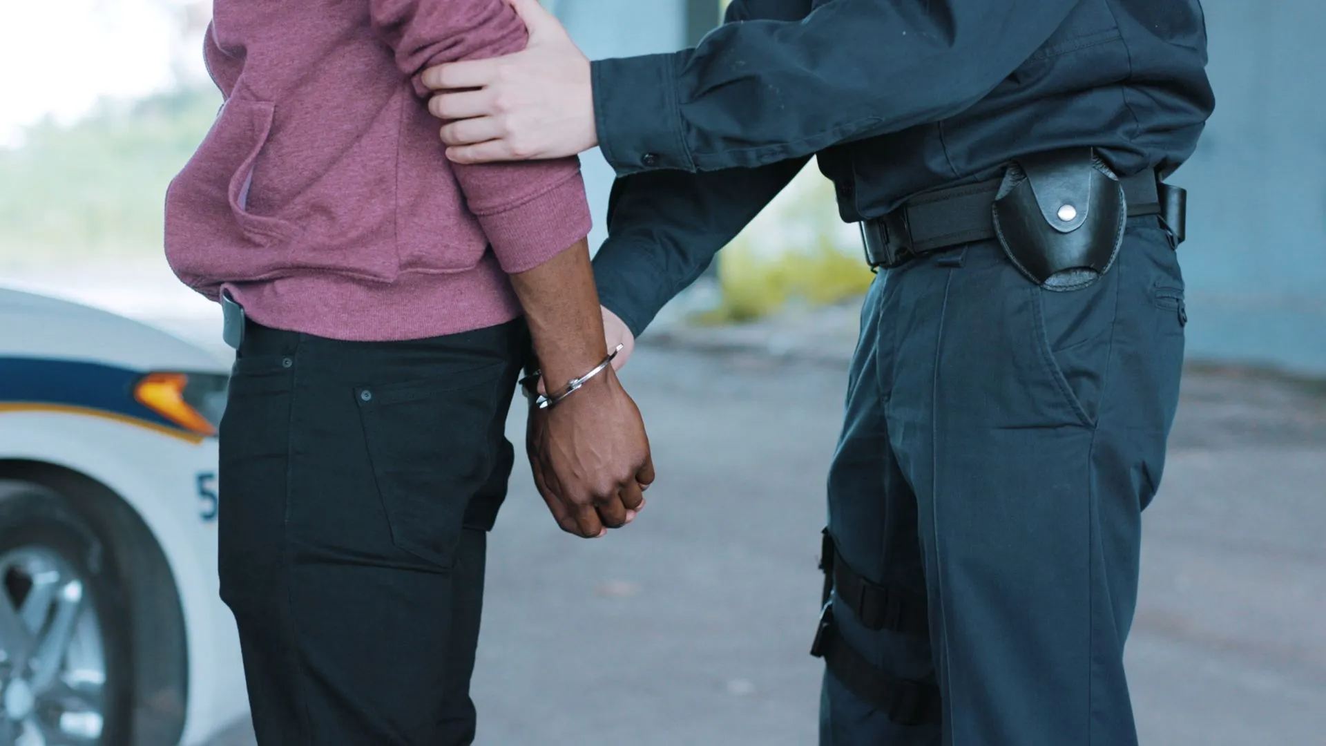 A police officer handcuffs a Black person next to a patrol car.