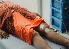 Persona acostada en una cama de hospital con vías intravenosas y monitores conectados a las piernas.