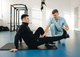 Dos personas haciendo ejercicio sobre colchonetas en un gimnasio luminoso; una ayuda a la otra con un estiramiento sentado.