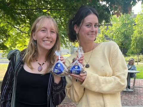 Two smiling individuals stand outdoors, each holding a small, clear bag of snacks with blue hamilton in front of them.
