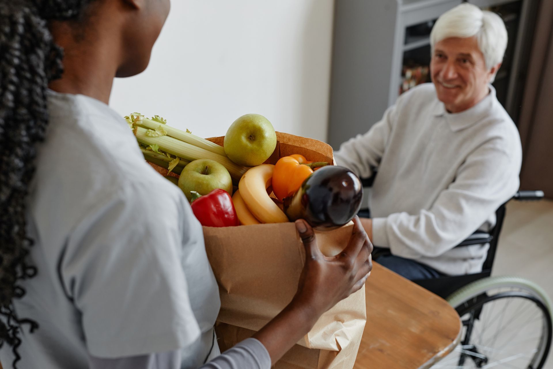 A woman is holding a bag of fruit and vegetables for an elderly man in a wheelchair.