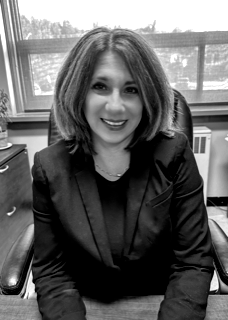 A woman in a suit is sitting at a desk in front of a window.