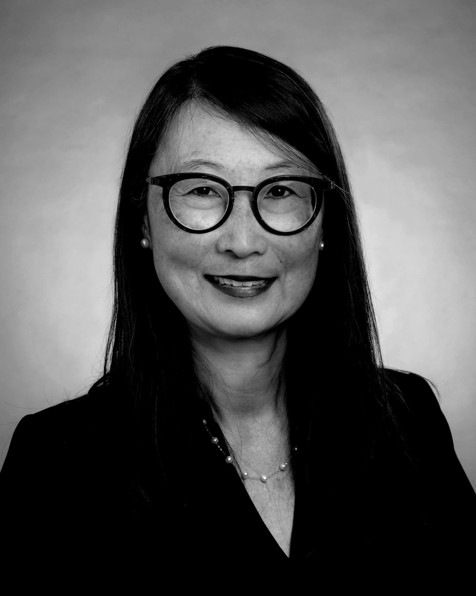 A woman wearing glasses and a black jacket is smiling in a black and white photo.