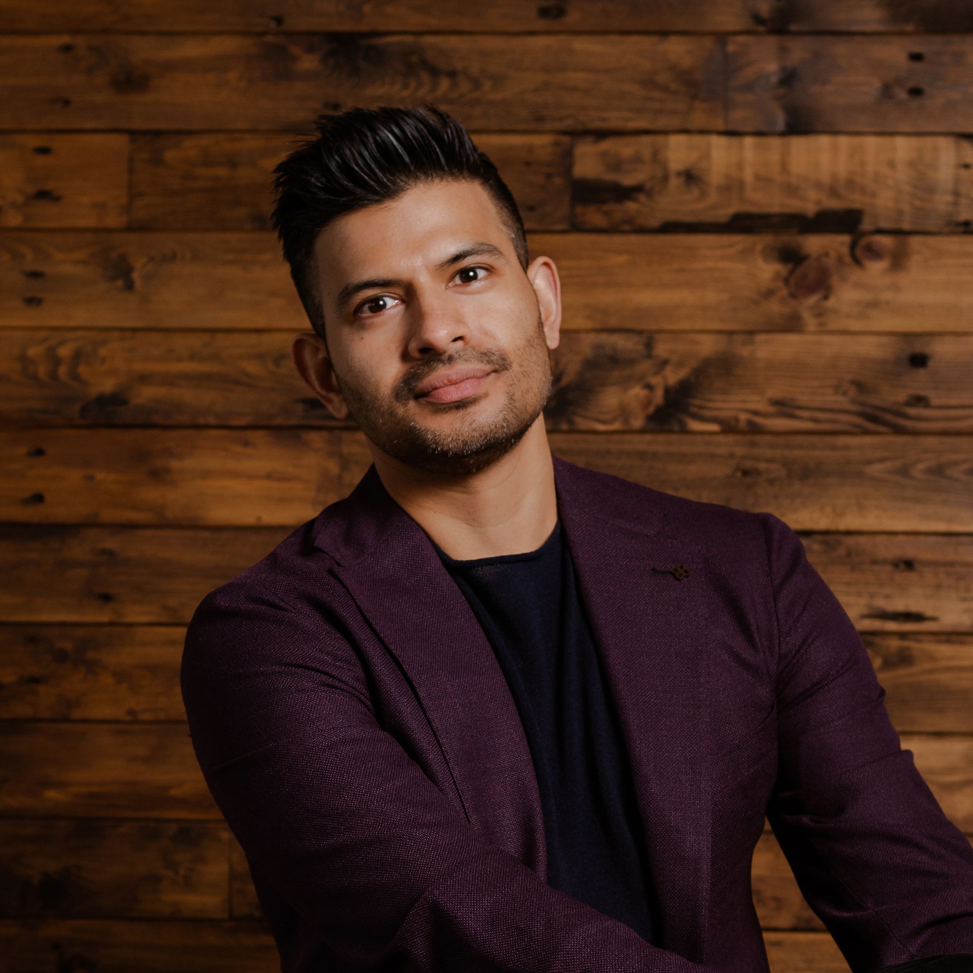 A man in a purple jacket and black shirt is sitting in front of a wooden wall.