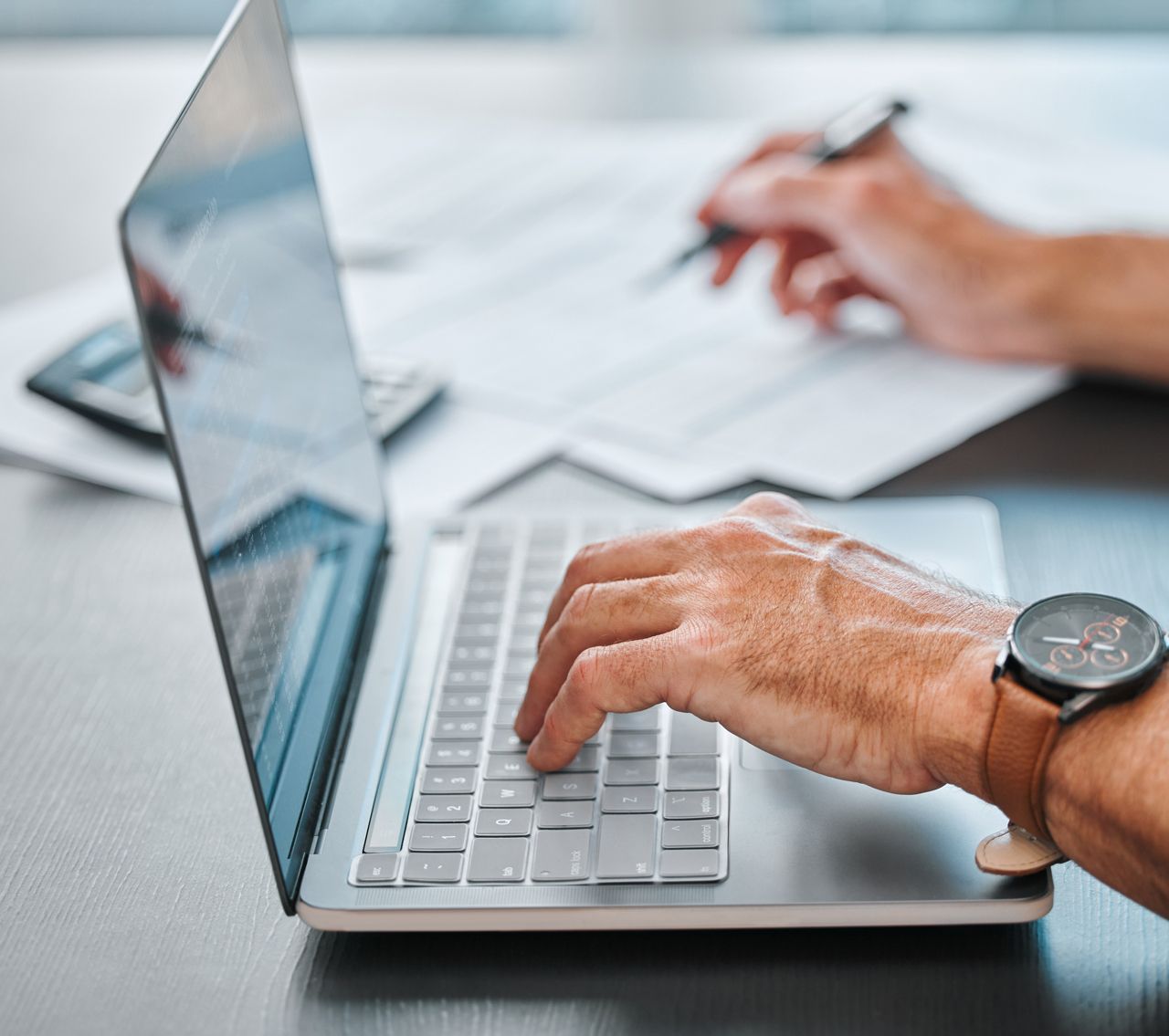 A person is typing on a laptop computer while writing on a piece of paper.