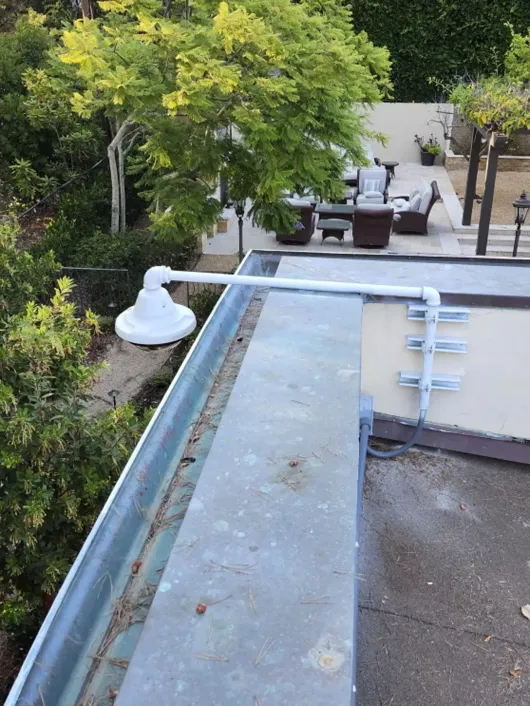 Rooftop terrace with a low wall, pipes, and a white fixture, overlooking a patio with trees and outdoor seating