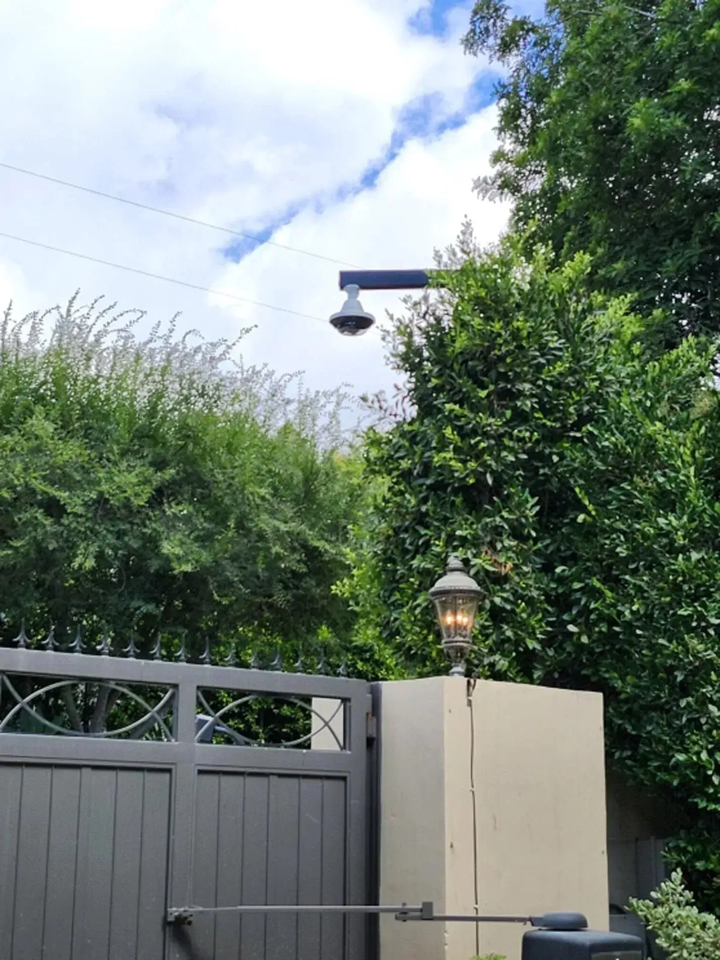 Gray gate and beige wall with lantern, dense green hedges, and a security camera against a cloudy sky.