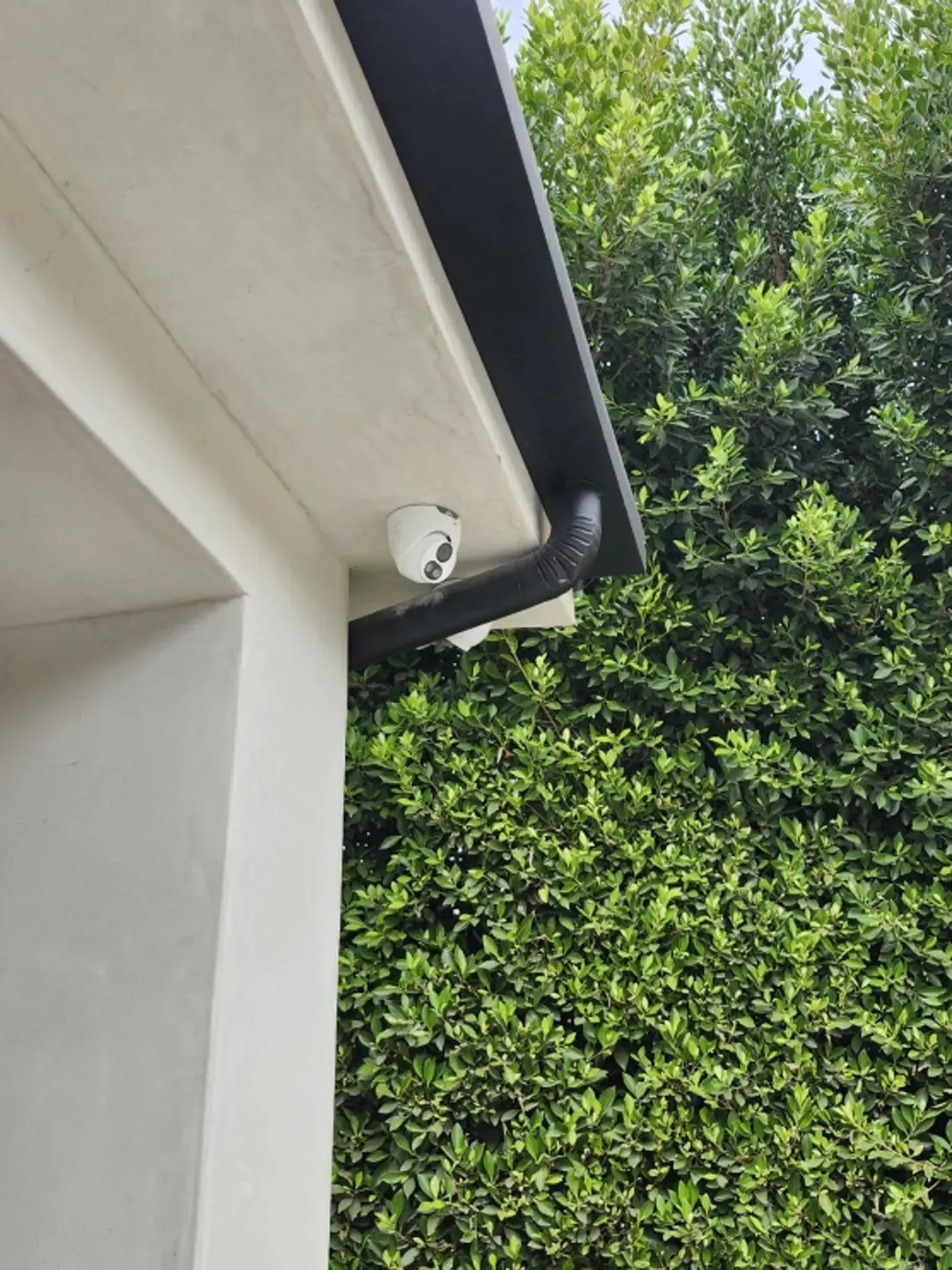 Outdoor corner of a house with a white security camera under the eaves beside a green hedge
