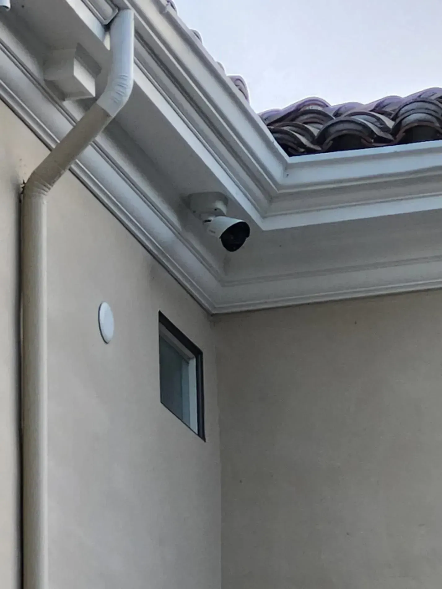 House exterior with beige stucco wall, roof eaves, gutter, window, and security camera.