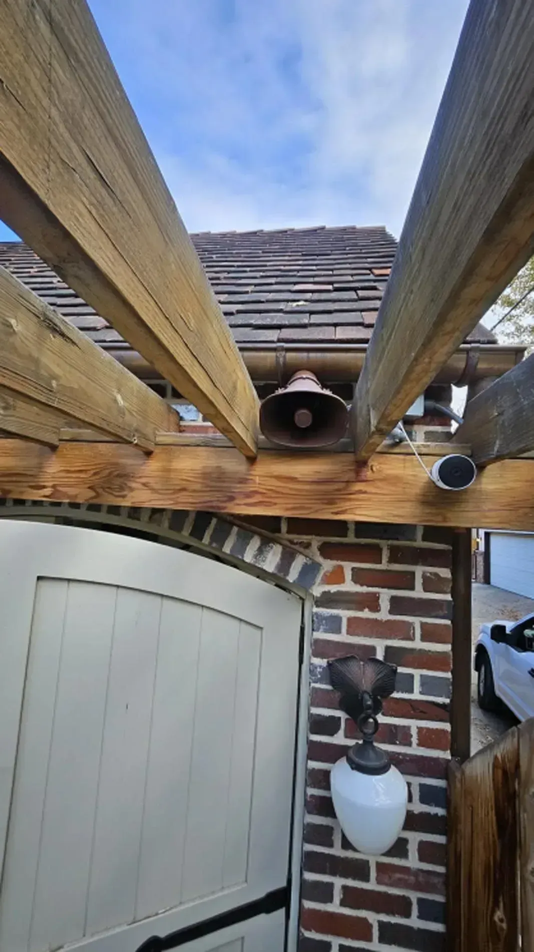 Under a wooden pergola, a ceiling fan and light are mounted above a doorway and brick wall.