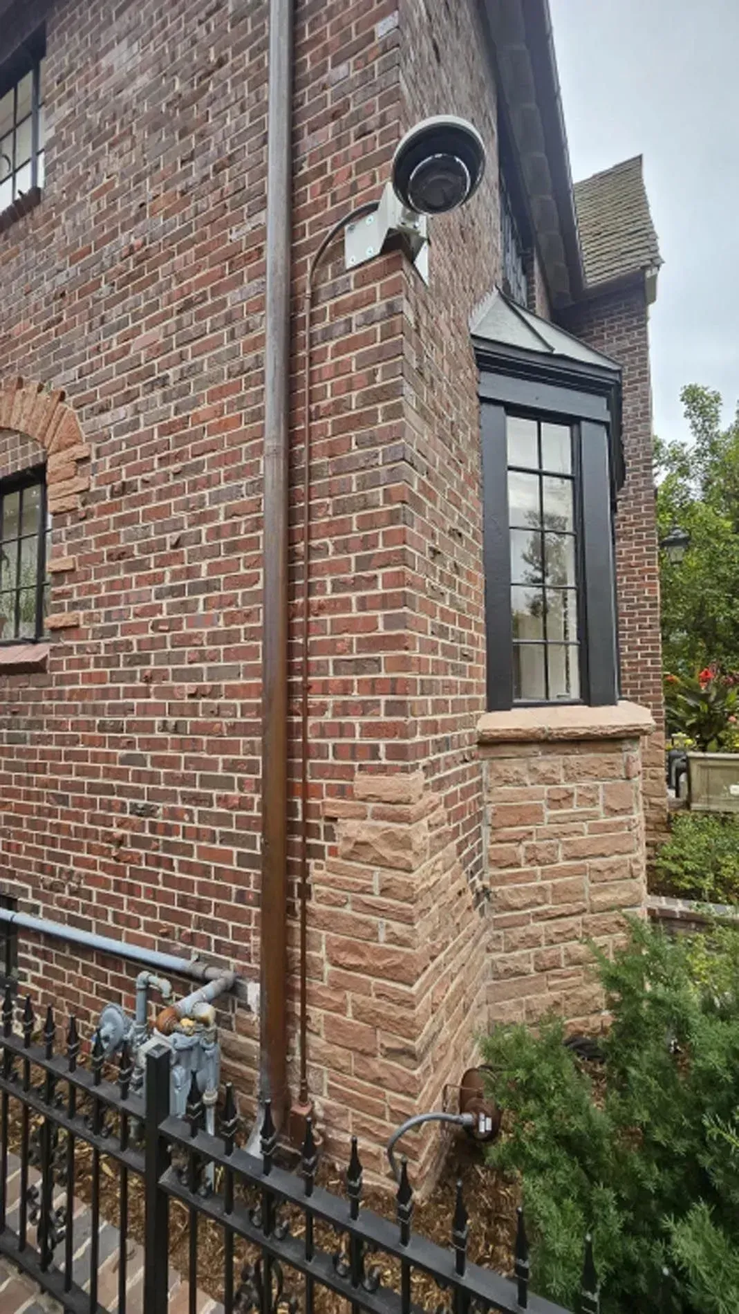 Brick house corner with black-framed bay window, stone base, and wrought-iron fence in front