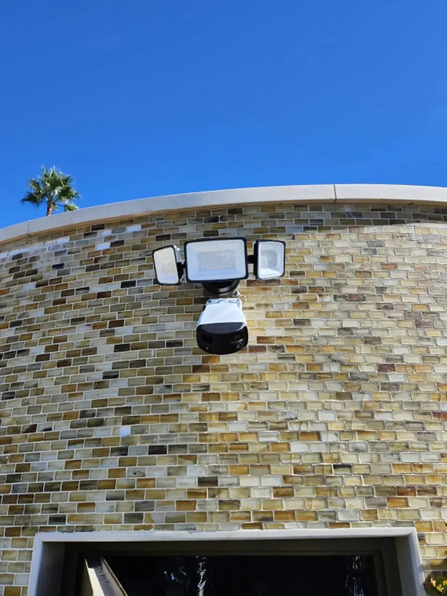 Wall-mounted security camera with lights on a stone building under a blue sky