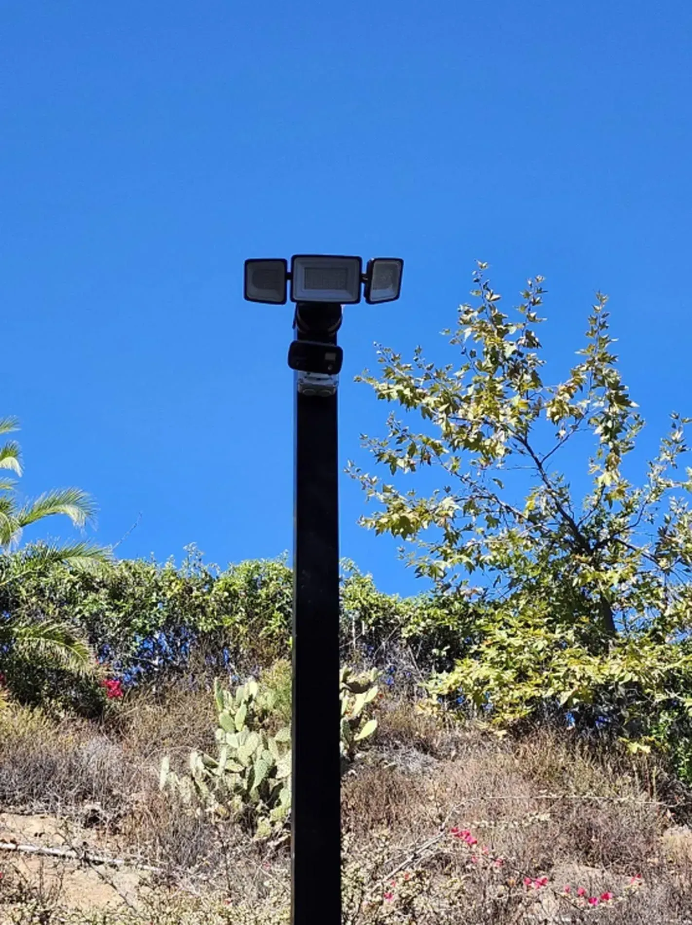 Trail camera on a black pole against blue sky and dry hillside vegetation