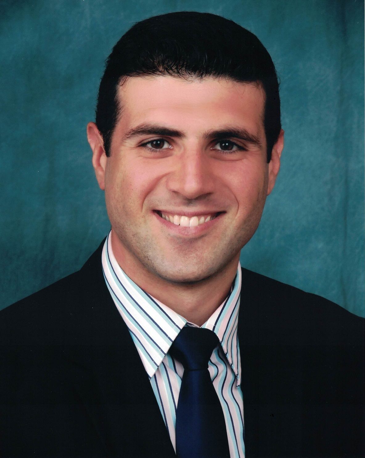 Man in a suit smiles, dark hair, blue and white striped shirt, and navy blue tie against a teal background.