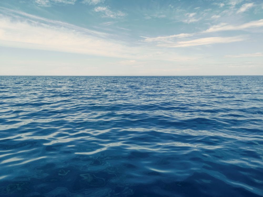 Calm, blue ocean meets a light blue, cloudy sky at the horizon.