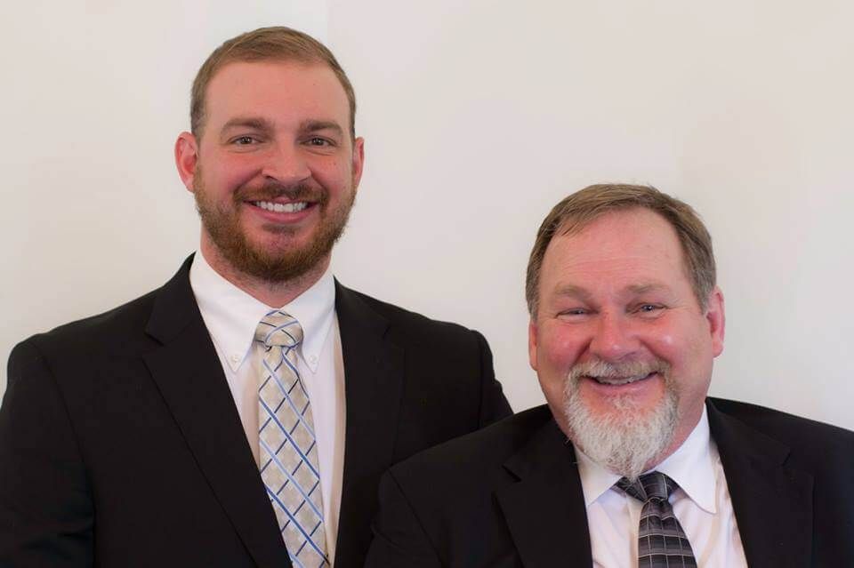 Two smiling men in suits standing against a plain light background