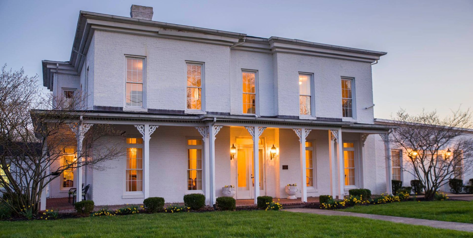 Two-story white farmhouse with lit windows and wraparound porch at dusk