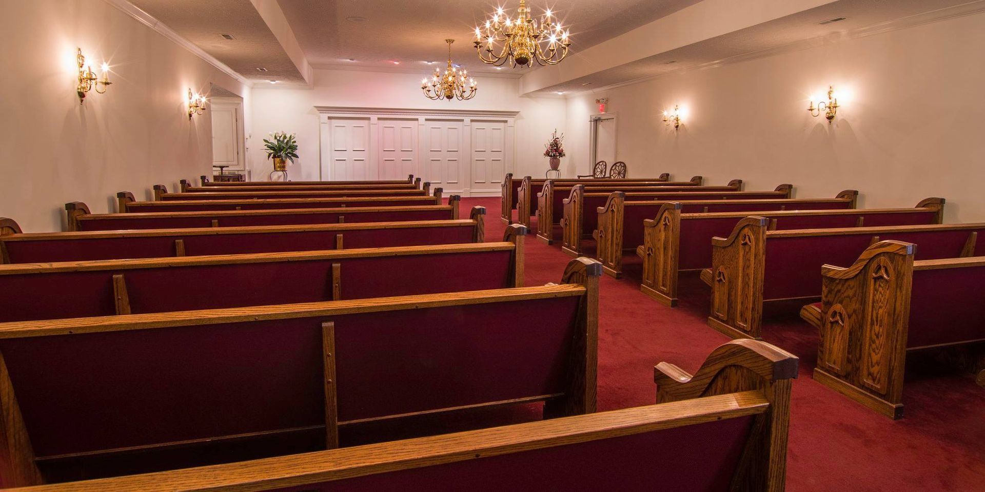 Empty church sanctuary with rows of red pews, white walls, and chandeliers under soft lighting