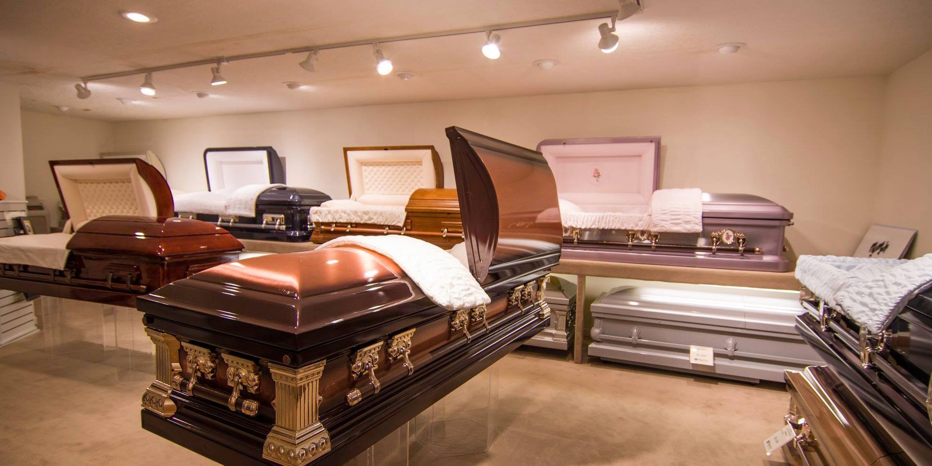 Caskets displayed in a funeral home showroom with beige walls and overhead lights