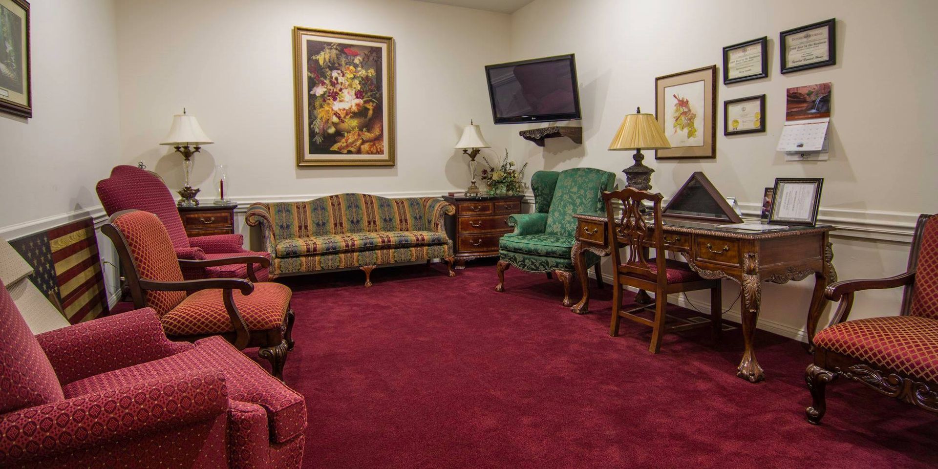 Cozy living room with burgundy carpet, patterned sofas, wall art, lamp, and a piano desk by the window.