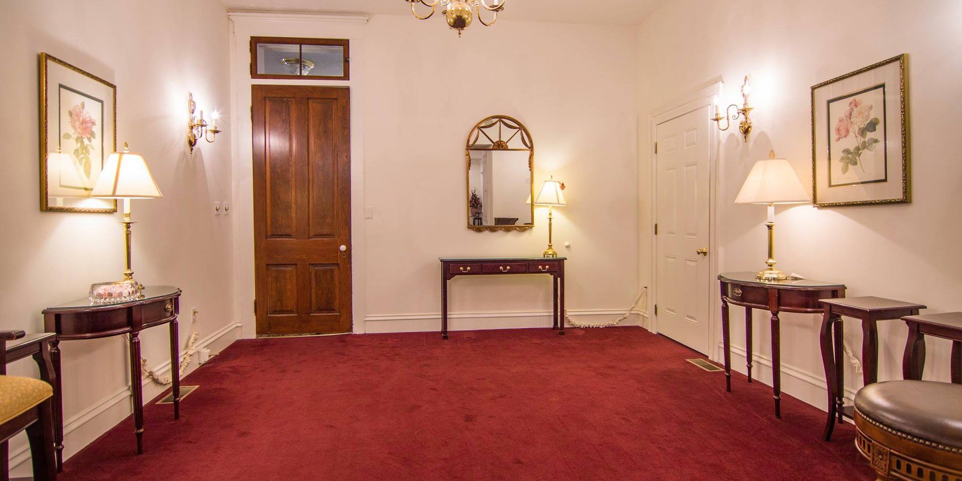 Elegant hallway with red carpet, wooden doors, wall sconces, and side tables with lamps and mirrors