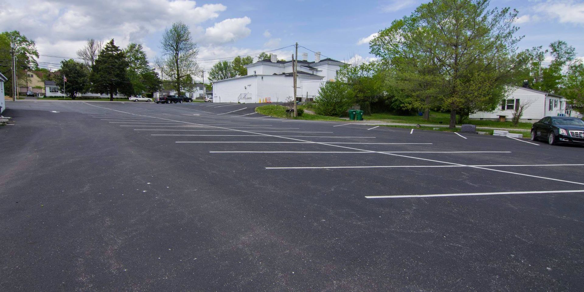 Empty parking lot with white lines, trees, and low buildings under a partly cloudy sky