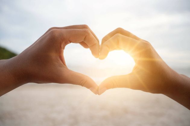Hands forming a heart shape around bright sunlight in a sky background