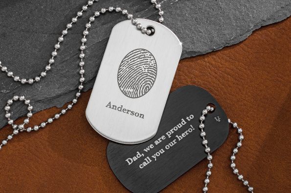 Two dog tags with fingerprint design, one saying “Dad, I’m proud to call you my father” on a brown fabric background