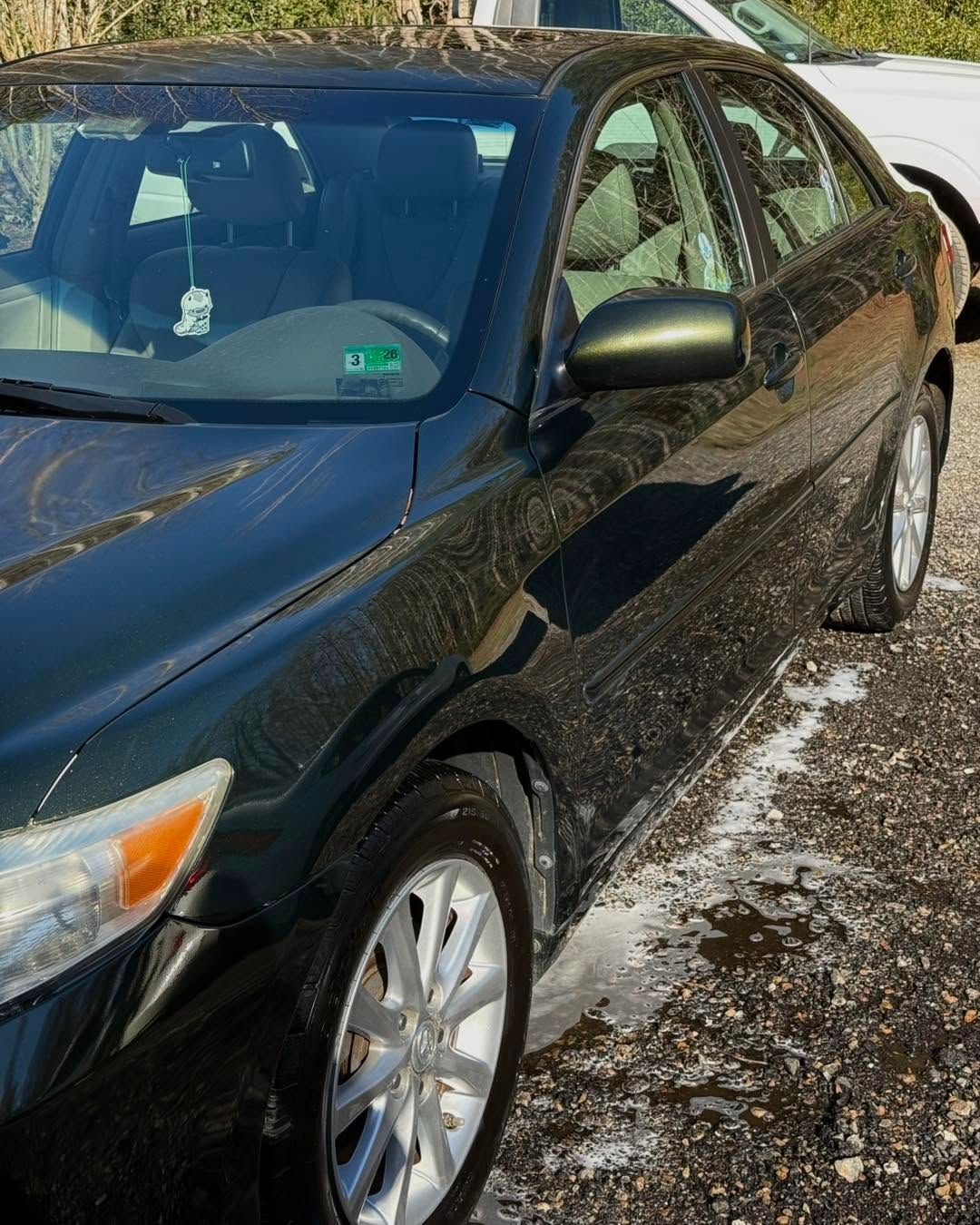 Black sedan parked on gravel in sunlight, front-left view.