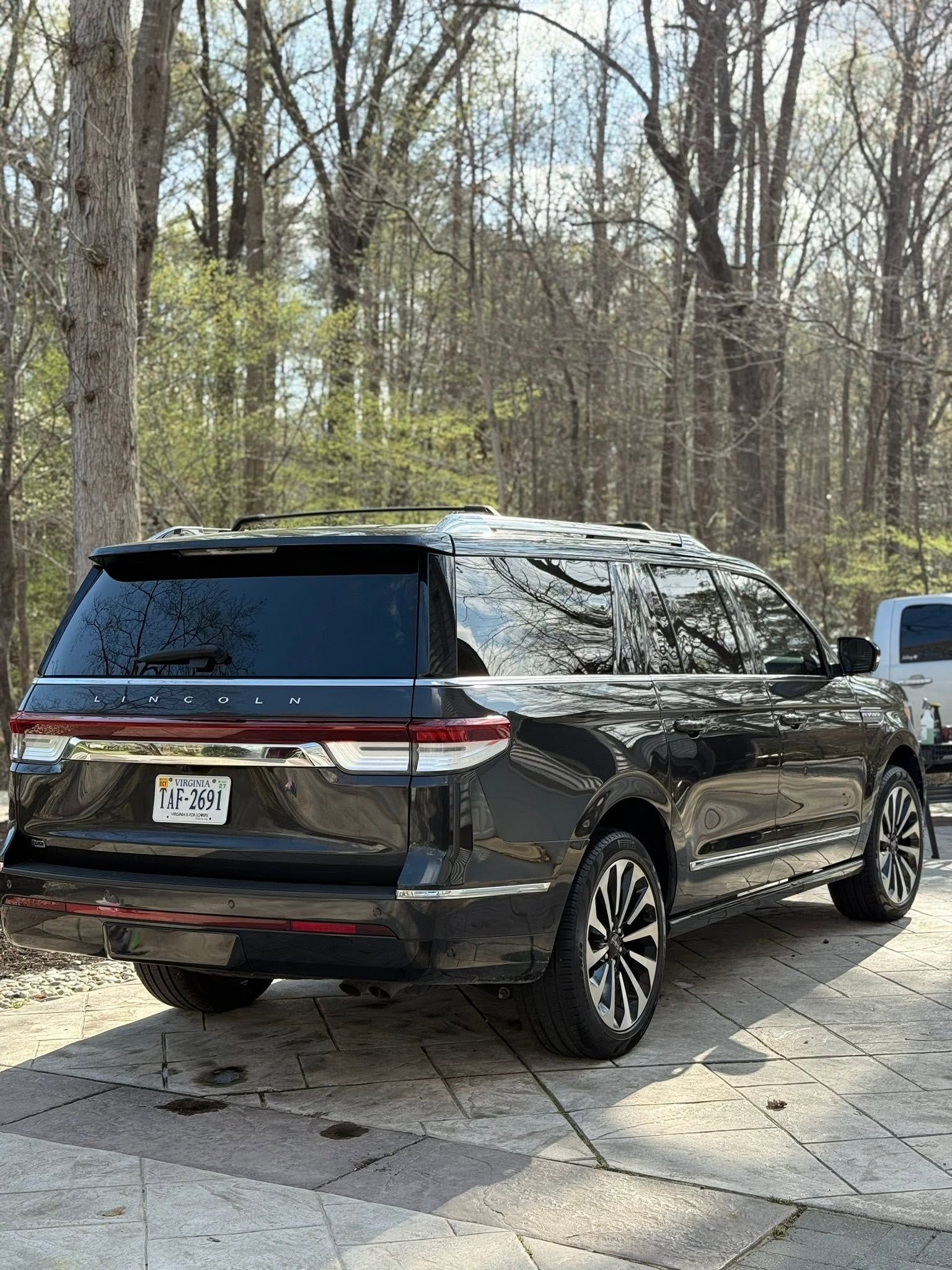 Black SUV parked on a driveway beside woods, with a tree reflected on its glossy rear window.