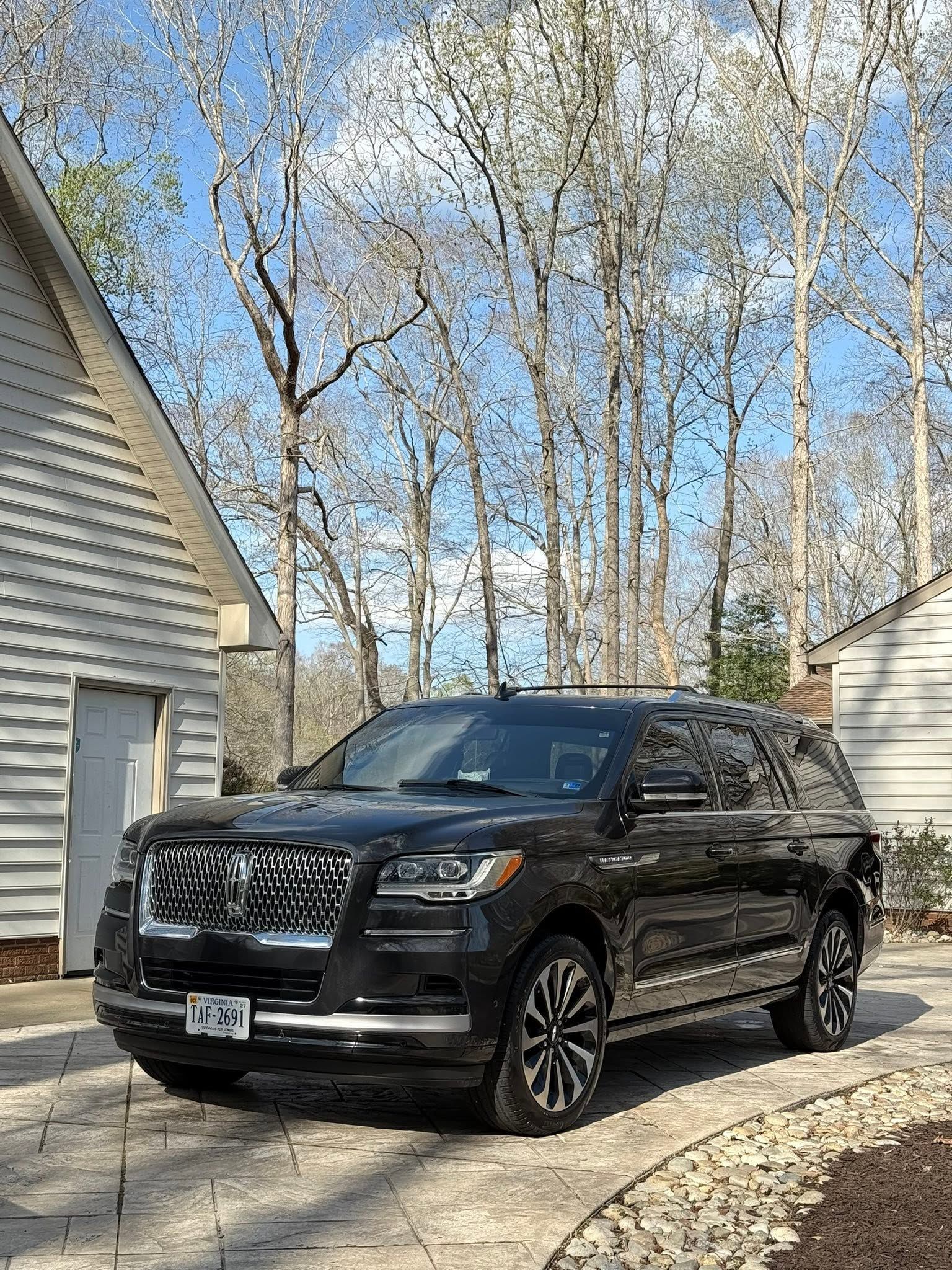 Black SUV parked in a driveway beside a house, with leafless trees and blue sky in the background.