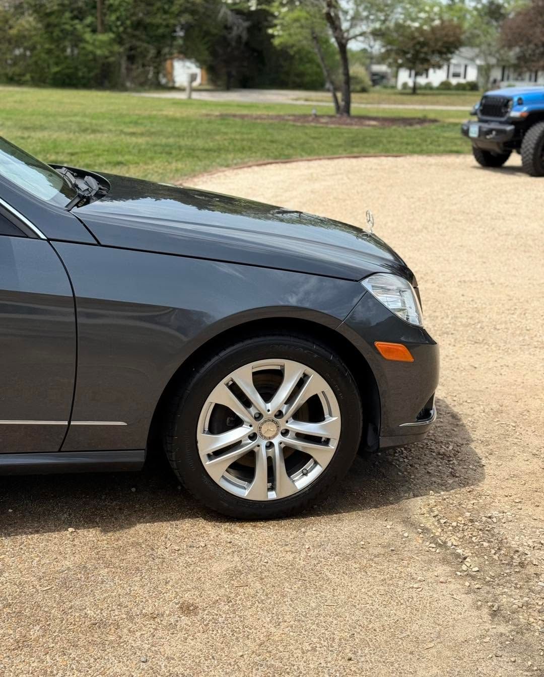 Front-left view of a dark gray sedan parked on a driveway near a yard, with a blue truck in the background.