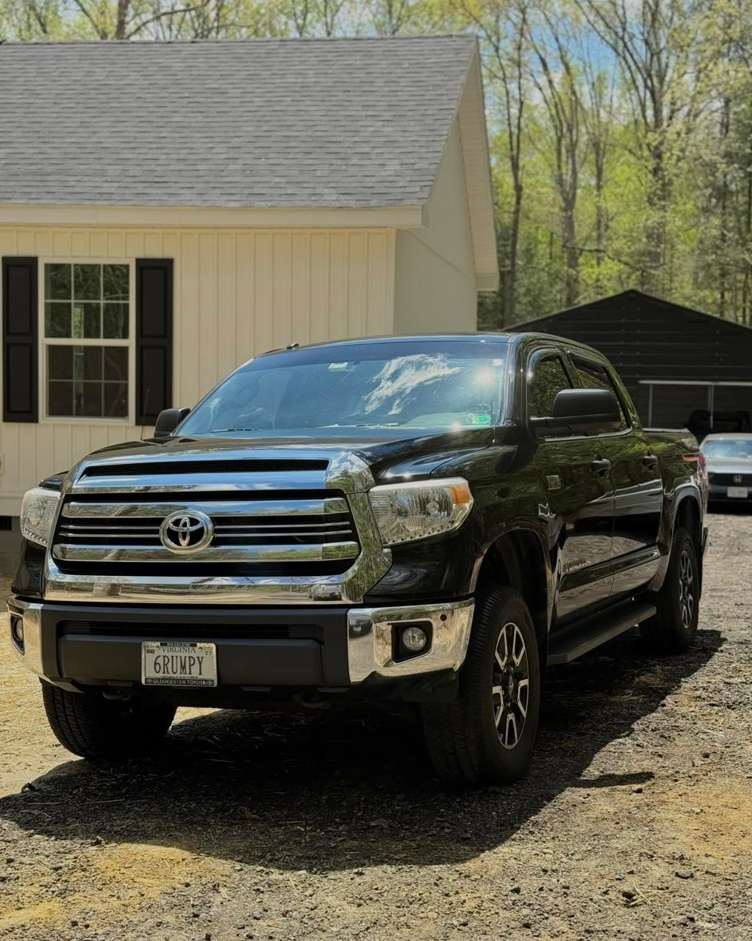 Black Toyota pickup truck parked on a gravel driveway beside a beige house and black shed.