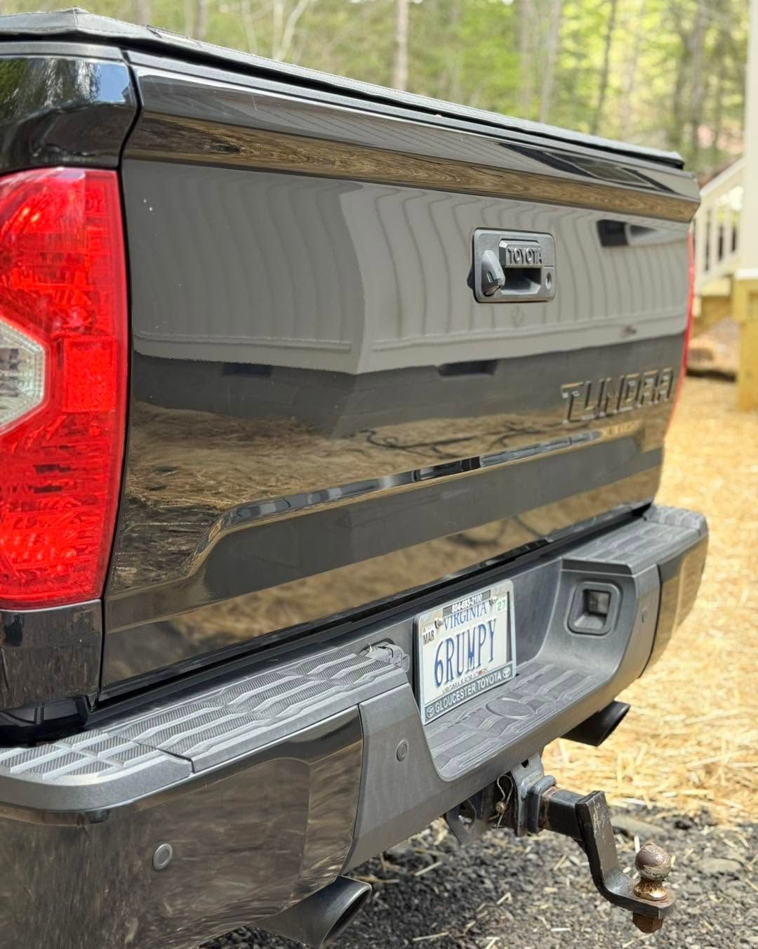 Rear of a black GMC pickup truck with tailgate, bumper, hitch, and license plate in a wooded driveway