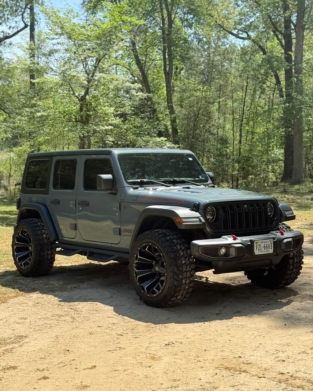 Gray Jeep Wrangler parked on a dirt path in a wooded area, with off-road tires and a front winch.
