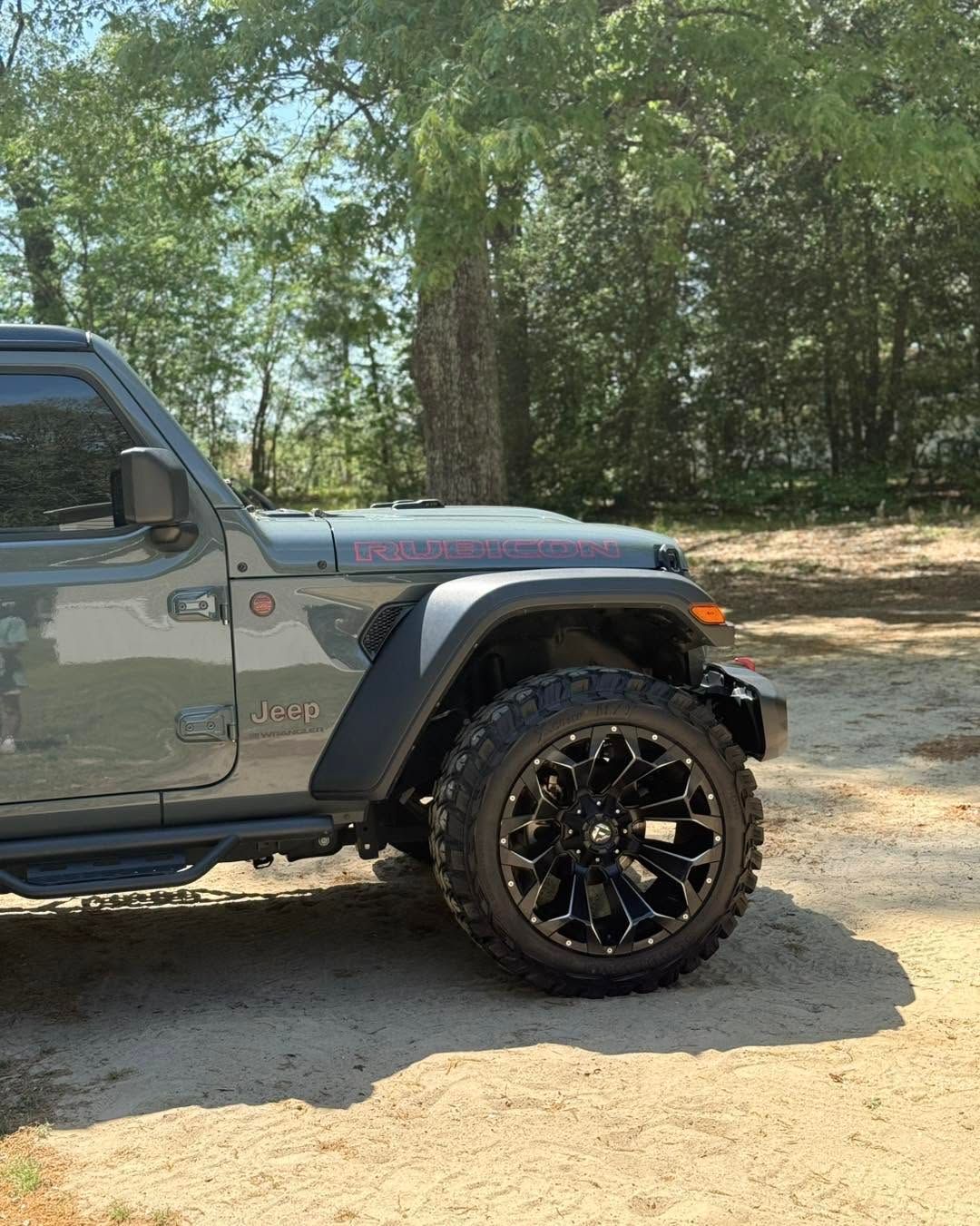 Gray off-road Jeep with black wheels parked on a dirt road by trees