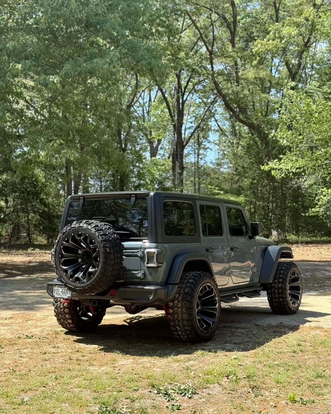 Gray Jeep Wrangler parked on a dirt path in a wooded area, rear view with spare tire.