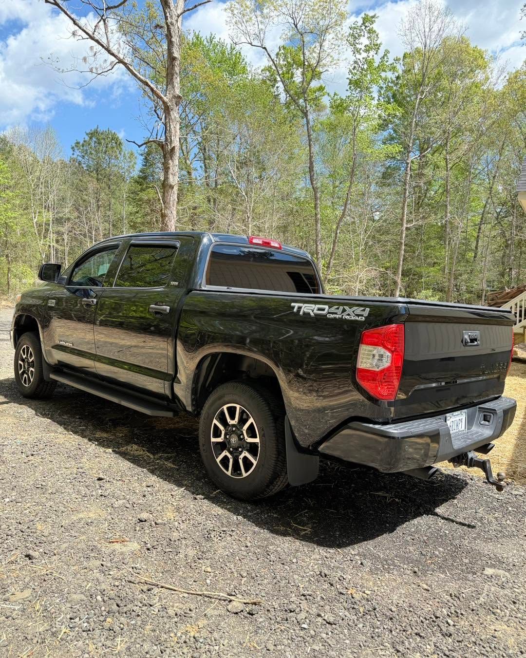 Black pickup truck parked on gravel by a wooded roadside under a blue sky