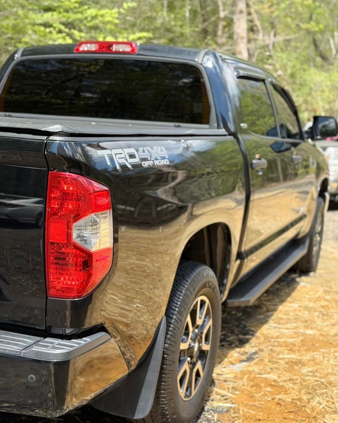 Black pickup truck parked on a dirt road in a wooded area, rear-left view