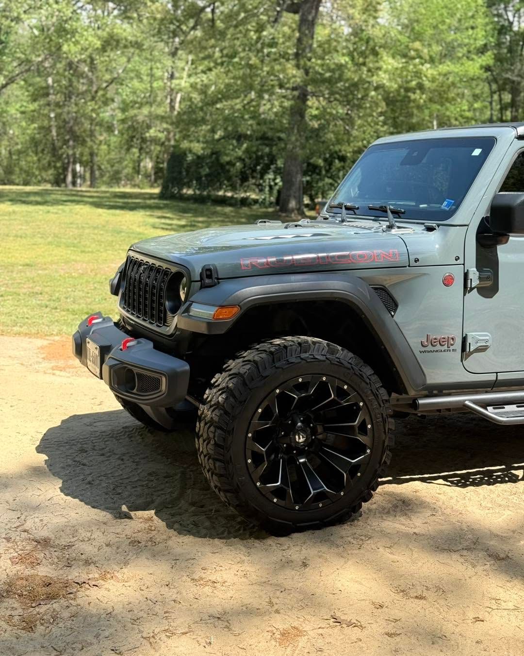 Black Jeep Wrangler with lifted tires parked on a dirt path in a sunny wooded area
