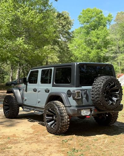 Gray lifted Jeep Wrangler parked on a dirt trail in a wooded area, with a spare tire on the back.