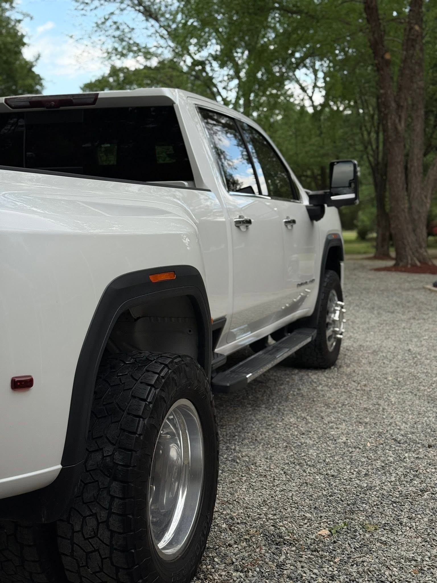 White lifted pickup truck parked on gravel beside trees and a road