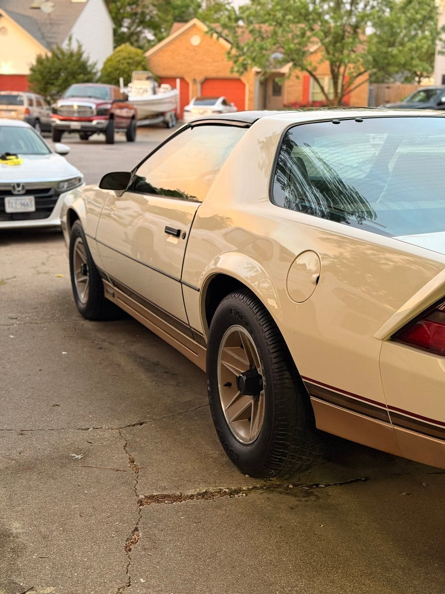 Beige classic car parked on a wet residential street, with houses and trees in the background.