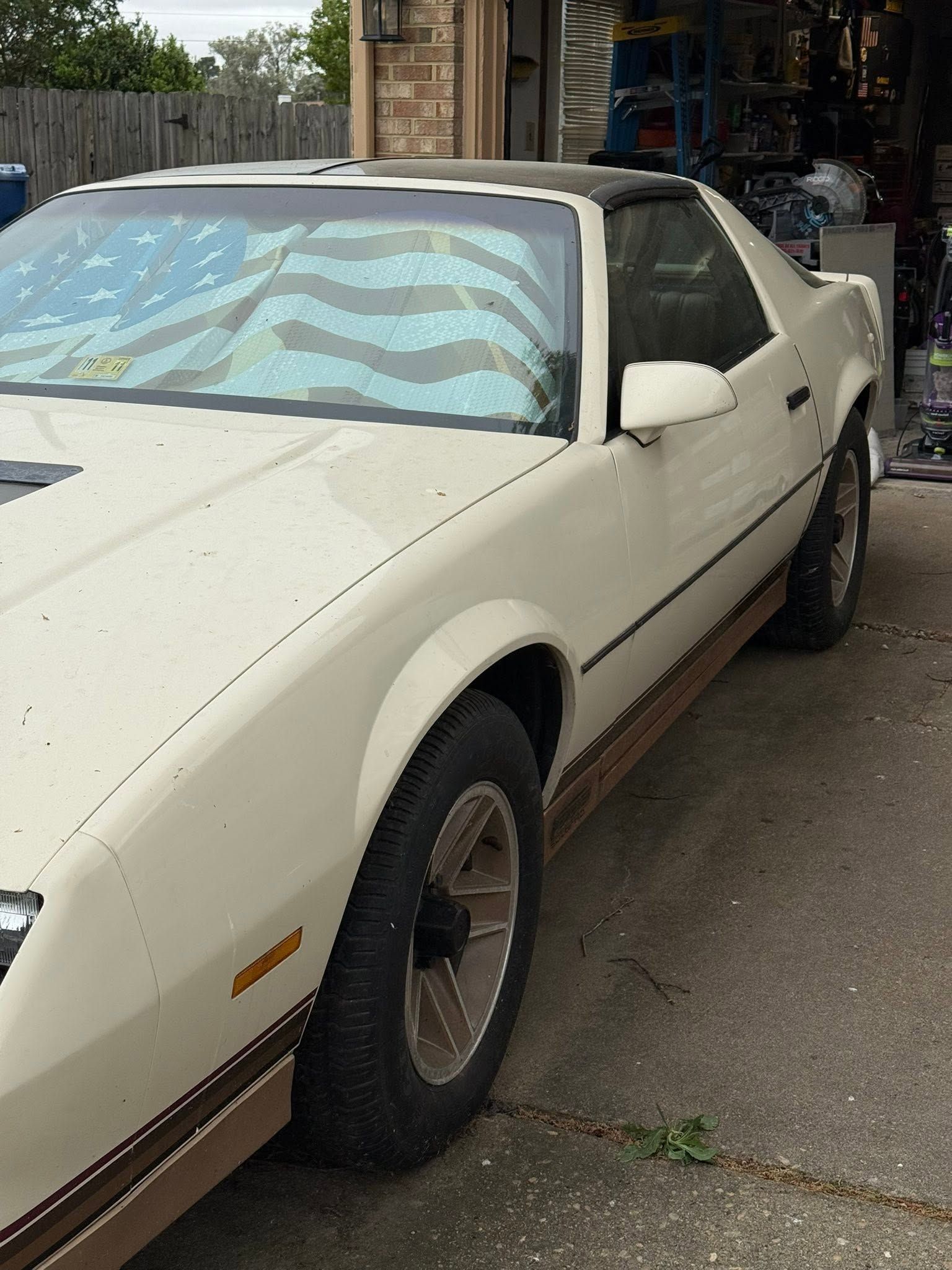White classic coupe parked outdoors beside a workshop, with a patterned sunshade in the windshield.