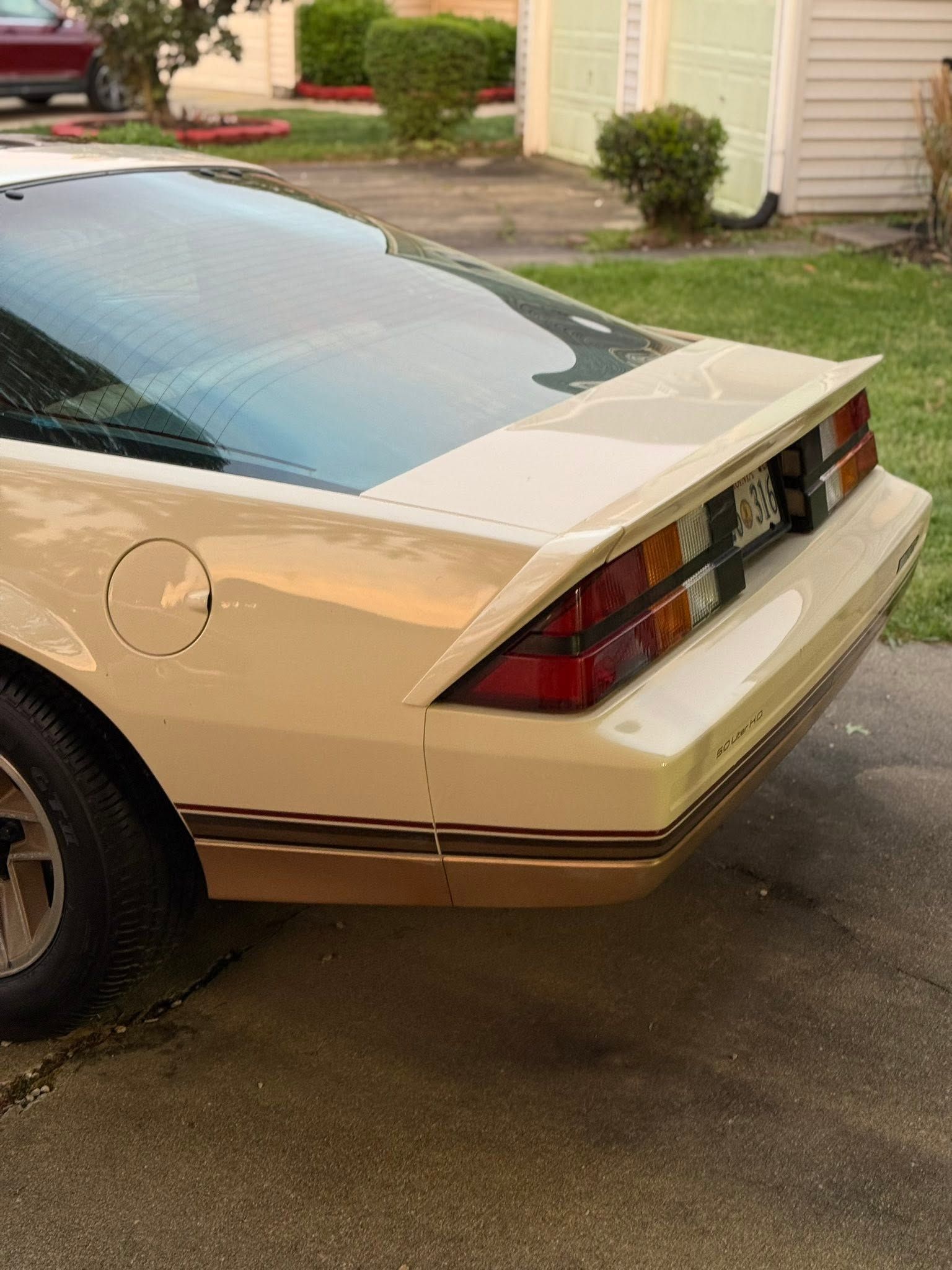White coupe parked in a driveway, rear view, with beige siding and shrubs in the background