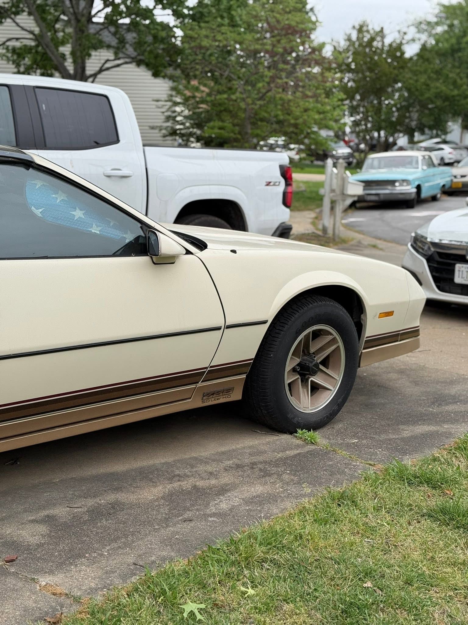 Cream-colored car parked beside a silver pickup in a lot, with trees and another car in the background.