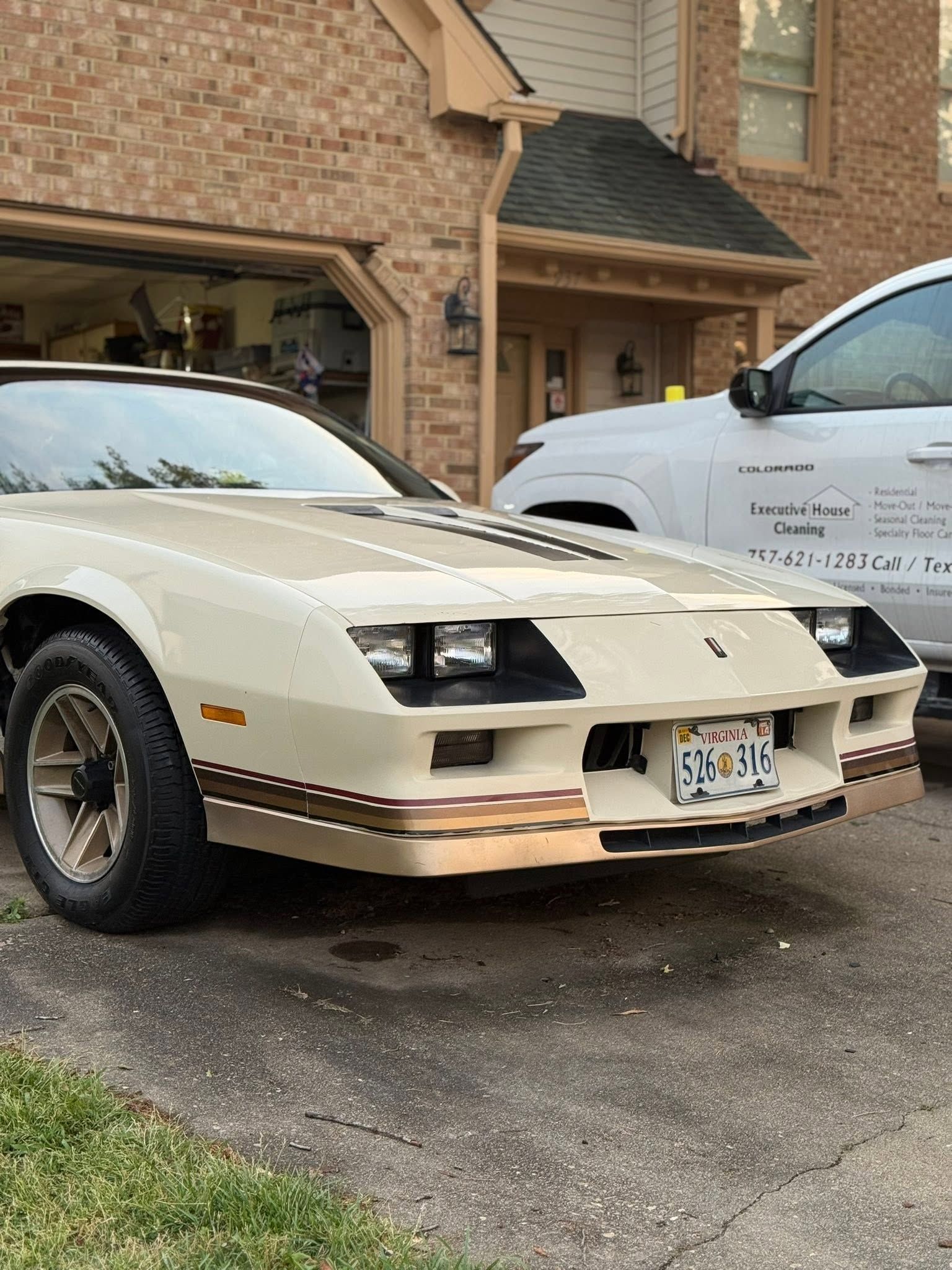 Cream-colored sports car parked in a driveway beside a white pickup in front of a brick house